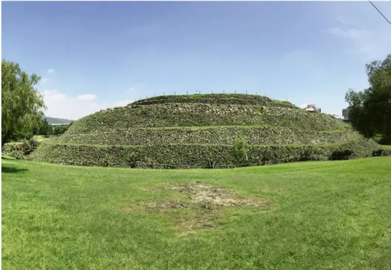 Fotografía de la pirámide circular de Cuicuilco, cubierta de vegetación, con escalinata y explanada verde.