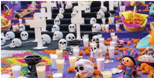 Fotografía de un altar (ofrenda) tradicional con calaveras blancas, velas encendidas, flores de cempasúchil, cruces de madera y una muñeca tradicional mexicana.