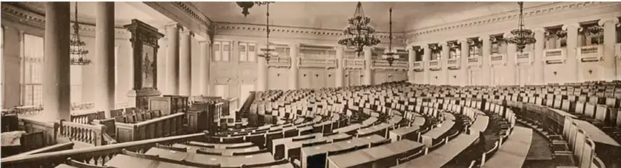 Fotografía en blanco y negro del interior de un salón parlamentario con curules dispuestas en semicírculo, columnas y candelabros, que representa la Duma Imperial de Rusia.