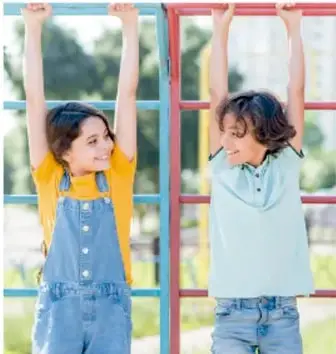 Fotografía de dos niñas colgadas de una estructura metálica de juegos, una con overol de mezclilla y otra con camiseta celeste.