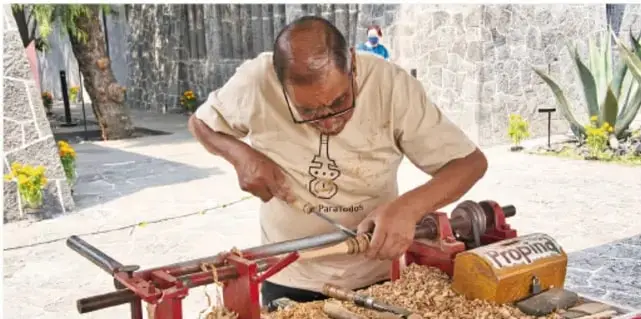 Artesano trabajando en un torno artesanal tallando madera, con virutas alrededor, en un entorno exterior con plantas.