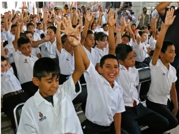 Fotografía de estudiantes de primaria en un aula levantando la mano para participar en una asamblea.