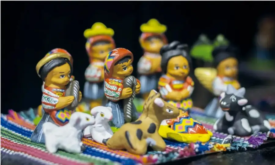 Fotografía de figuras artesanales de barro o cerámica pintadas a mano: personas con trajes tradicionales y sombreros, junto con animales (ovejas, un toro, un perro), colocadas sobre un tejido de colores; son artesanías típicas de los mercados indígenas.