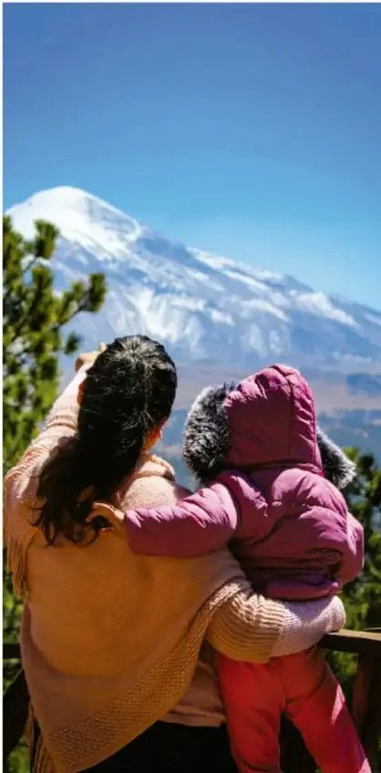 Fotografía de una madre de espaldas cargando a un niño, ambos contemplando un volcán nevado bajo un cielo azul, con árboles de pino a los lados; evoca la relación de las comunidades con su entorno natural.