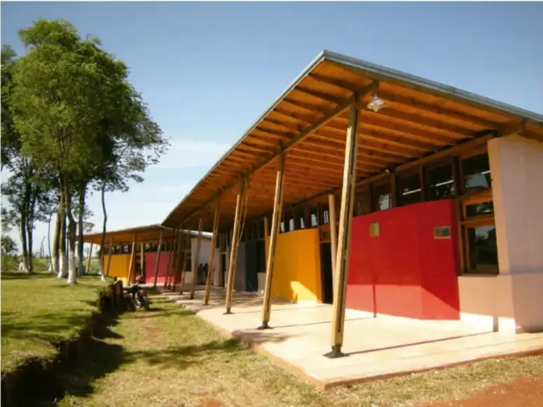 Fotografía de un edificio escolar rural de una sola planta, con techo de lámina sostenido por pilares de madera, muros pintados de rojo, amarillo y blanco, y un patio de tierra con árboles y pasto alrededor.