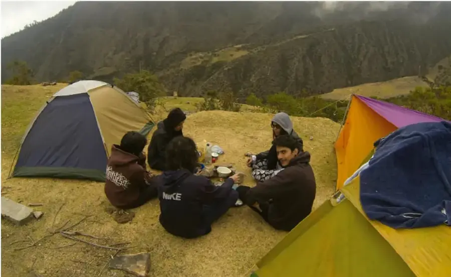 Grupo de jóvenes acampando en una zona boscosa de montaña, sentados junto a tiendas de campaña, algunas personas preparando alimentos.