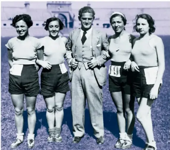 Fotografía histórica con tinte violeta de un grupo de cuatro mujeres atletas en camisetas claras y shorts oscuros con números de competidor, junto a un hombre de traje, al aire libre frente a un edificio antiguo.