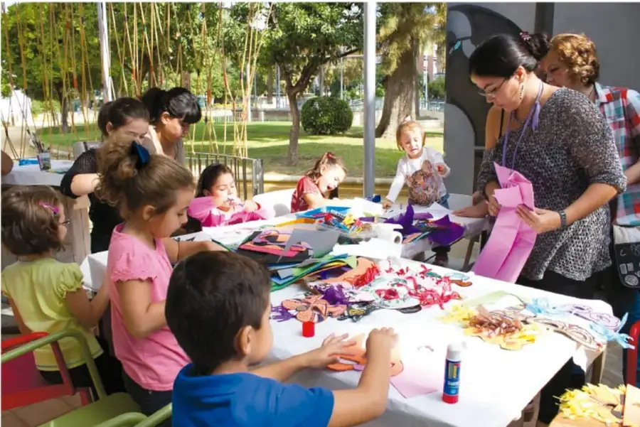 Fotografía de un grupo de niñas, niños y adultos trabajando en una mesa larga al aire libre, rodeados de árboles; recortan y pegan papel de colores con tijeras y colbón para crear obras de arte (posiblemente en un taller comunitario o escolar).
