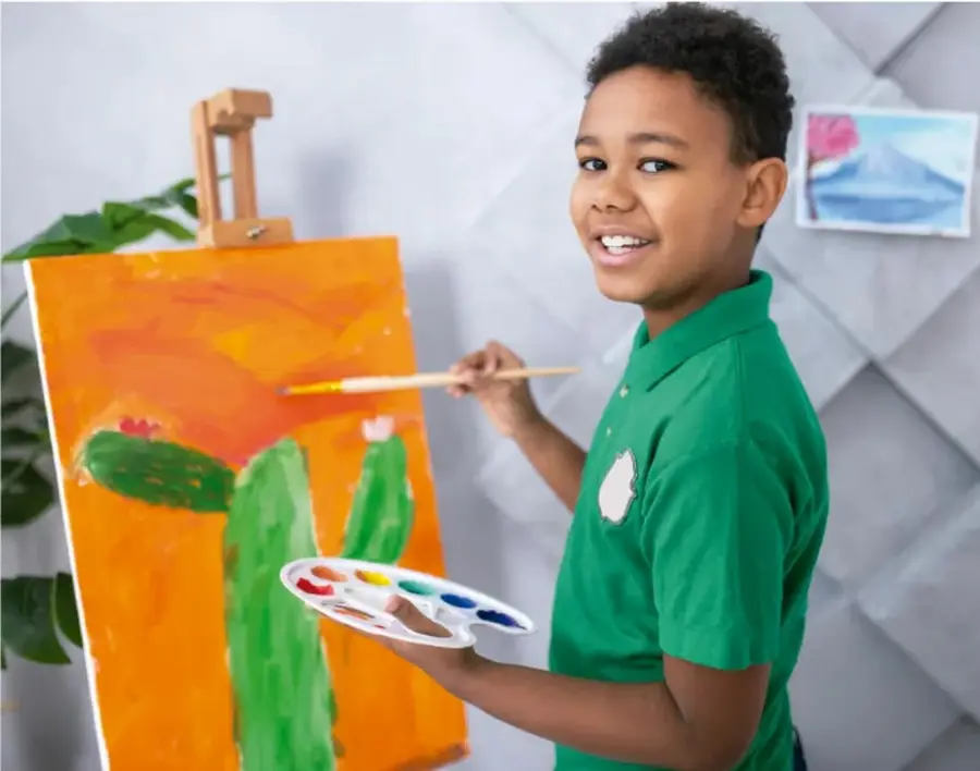 Niño con camisa verde pintando un lienzo naranja con cactus en un caballete, sosteniendo una paleta de colores, representando el descubrimiento de talentos.