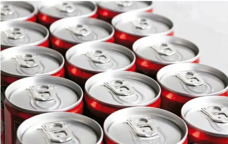 Fotografía cenital de múltiples latas de aluminio de refresco de cola, con sus anillas de apertura visibles, dispuestas en filas; ilustra el ejemplo del consumo de recursos en la producción de refrescos.