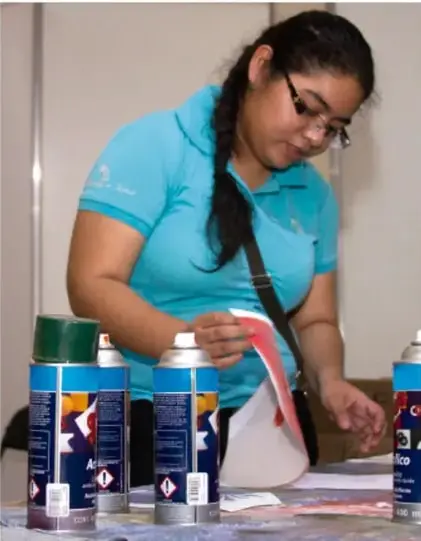 Fotografía de una mujer joven con playera azul y coleta, sosteniendo un bote de aerosol frente a una mesa con materiales.
