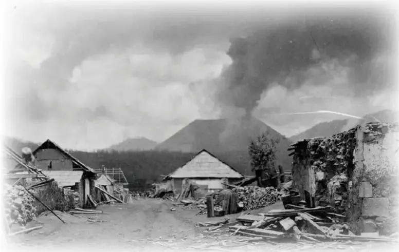 Fotografía en blanco y negro que muestra el volcán Paricutín en erupción, con una nube de ceniza, y ruinas de edificios del pueblo de San Juan Parangaricutiro en primer plano.