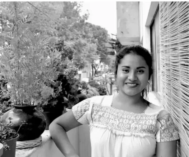 Fotografía en blanco y negro de una joven mexicana sonriente, con cabello recogido y blusa bordada, posando junto a una pared y plantas.