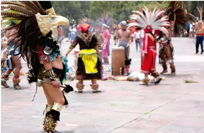 Grupo de personas con trajes, máscaras y penachos tradicionales danzando en una plaza al aire libre; representan una actividad artística expresiva comunitaria (danza tradicional).