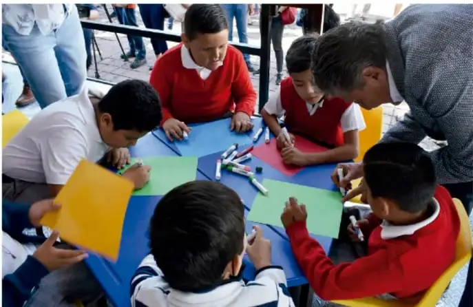 Fotografía de un grupo de estudiantes de primaria trabajando en una mesa con hojas de colores y marcadores, acompañados por un maestro, mientras elaboran un proyecto escolar.