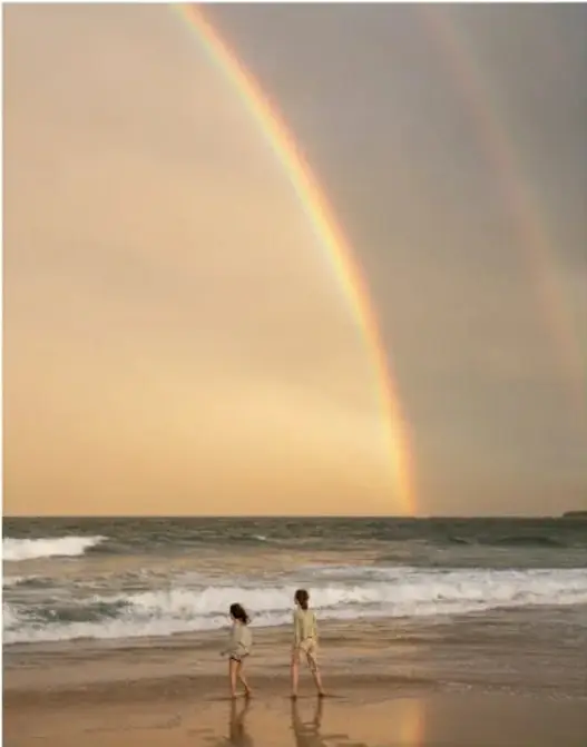 Fotografía de dos niños pequeños caminando en la playa bajo un arcoíris doble, con el mar y el cielo al fondo.