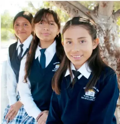 Fotografía de tres estudiantes con uniforme escolar (chamarra azul marino con escudo, camisa blanca y corbata) posando bajo un árbol, en el patio de la escuela.
