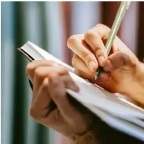 Fotografía de las manos de una persona escribiendo con un lápiz sobre un cuaderno.