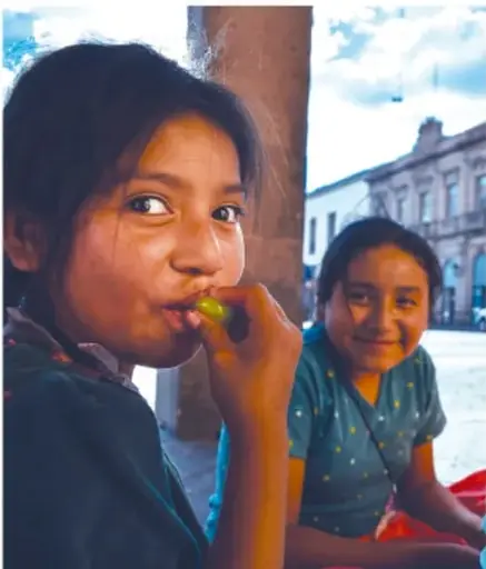 Fotografía de dos niñas indígenas sonriendo; una de ellas come una fruta verde. Al fondo se observa una calle con edificios.