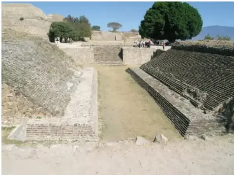 Fotografía de una cancha de juego de pelota mesoamericana, con sus taludes de piedra escalonados y montículos cercanos, al aire libre.