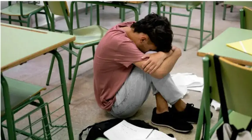Fotografía de un joven sentado en el piso de un salón de clases, con la cabeza entre las rodillas y los brazos abrazando las piernas, rodeado de pupitres verdes y papeles tirados en el suelo; transmite abatimiento o frustración.