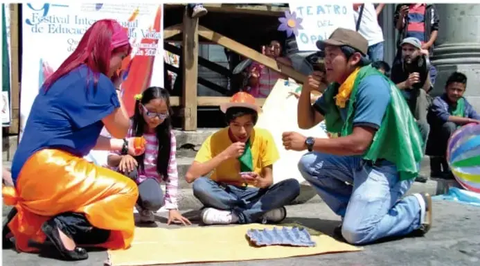 Fotografía de una representación de teatro del oprimido al aire libre. Una mujer con cabello rojo y vestimenta azul y naranja interactúa con niños y jóvenes sentados en el suelo. Un joven con gorra y pañuelo verde sostiene un micrófono. Detrás hay carteles que dicen 'Festival Internacional de Educación' y 'TEATRO DEL OPRIMIDO'. Los participantes están sobre una manta amarilla.