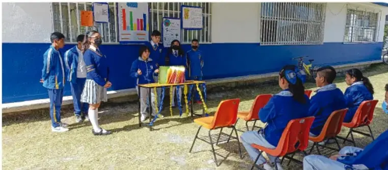Fotografía de alumnos de Telesecundaria presentando un proyecto en el patio de su escuela ante compañeros sentados en sillas; incluye gráficas y carteles en la pared.