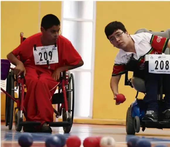 Fotografía de dos deportistas en silla de ruedas (dorsales 209 y 208) practicando parapan (boccia) en una cancha techada, lanzando esferas rojas y azules.