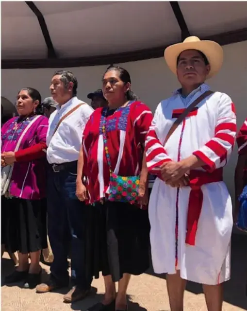 Fotografía de integrantes de una comunidad indígena durante la ceremonia de cambio de poderes: una mujer con huipil bordado, un hombre con camisa y cinta, otra mujer con traje tradicional y un hombre con sombrero de ala ancha y túnica blanca con cinta roja.