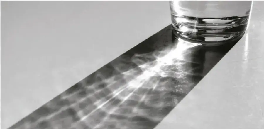 Fotografía en blanco y negro de un vaso con agua sobre una superficie; la luz se refracta y se proyecta una franja luminosa con textura ondulada.