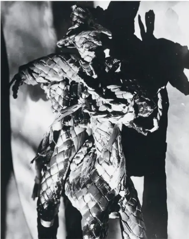Fotografía en blanco y negro de una figura humana cubierta con materiales reflectantes, con una sombra marcada sobre una pared.