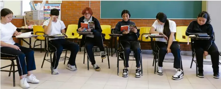 Fotografía de seis estudiantes sentados en sillas con pupitre en el aula, leyendo libros y preparando material de trabajo.