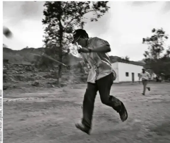 Fotografía en blanco y negro de un hombre ralamul corriendo por un camino rural mientras patea la bola; al fondo se ven cerros, árboles y un edificio pequeño. Crédito: Francisco de la Luz Martínez Álvarez.