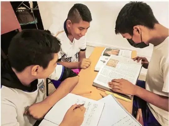 Fotografía de tres estudiantes de telesecundaria trabajando juntos en una mesa: uno escribe en un cuaderno, otro revisa un libro abierto y el tercero observa; llevan cubrebocas. Crédito: Luis Eduardo García Mural, Tlaxiaco.