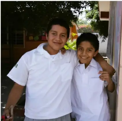 Fotografía de dos estudiantes varones con uniforme blanco, sonriendo y abrazándose, en un espacio escolar con vegetación.