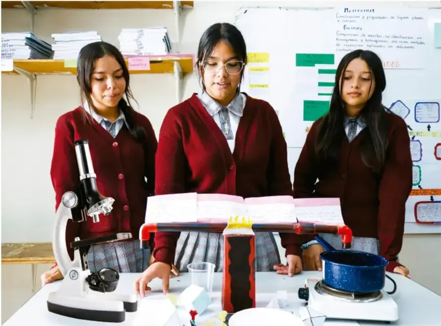 Fotografía de tres estudiantes de telesecundaria con uniforme, junto a una mesa con microscopio, utensilios de laboratorio y un modelo artesanal, preparando materiales de su proyecto.