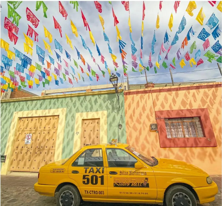 Calle adorned con papal picado multicolor cruzado sobre fachadas de colores y un taxi amarillo numerado 501 estacionado frente a un portón.
