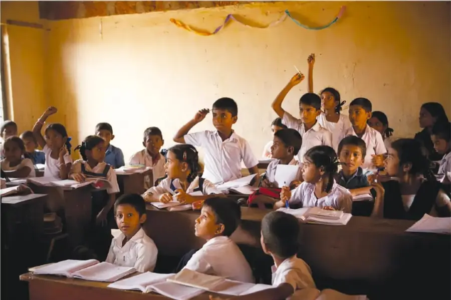 Fotografía de un aula rural con estudiantes de primaria sentados en bancas de madera; varios levantan la mano y sonríen, con cuadernos sobre sus pupitres.