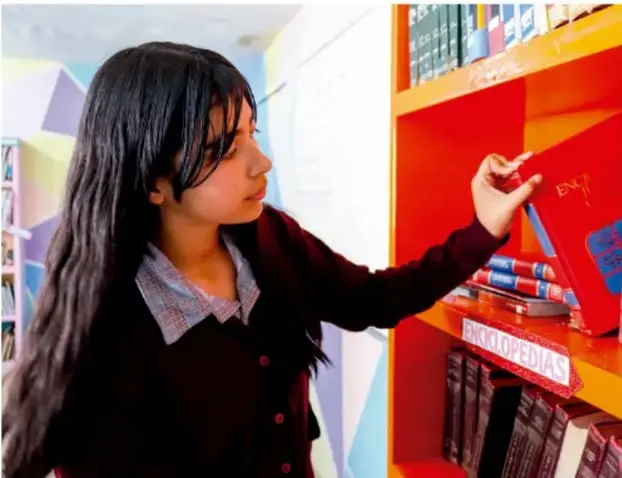 Fotografía de una estudiante junto a una estantería de biblioteca tomando o revisando un libro, ilustrando la investigación en fuentes.