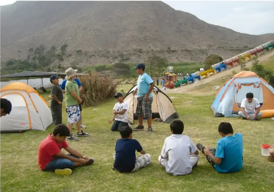 Fotografía de un grupo de adolescentes en un campamento al aire libre con tiendas de campaña, sentados y de pie sobre el pasto, con cerros y estructuras recreativas al fondo.