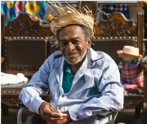 Fotografía de un hombre adulto mayor, probablemente de comunidad indígena, con corona o tocado de paja, sentado y sonriente, con textiles y una escultura de madera al fondo.