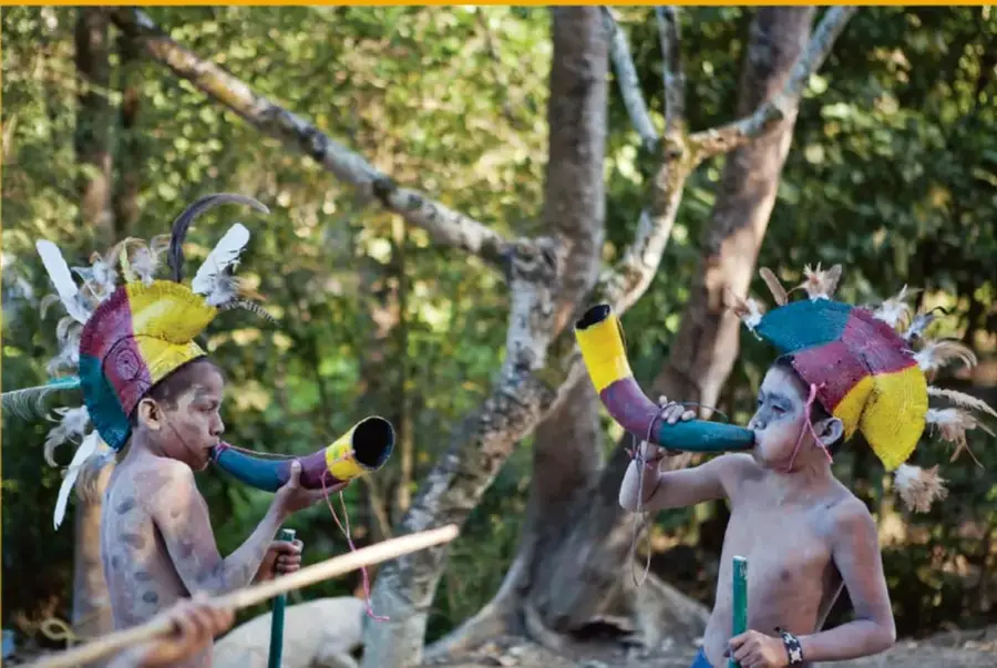 Fotografía de dos niños indígenas semidesnudos con penachos de plumas de colores, pintura corporal y trompetas o instrumentos de viento, en un bosque.