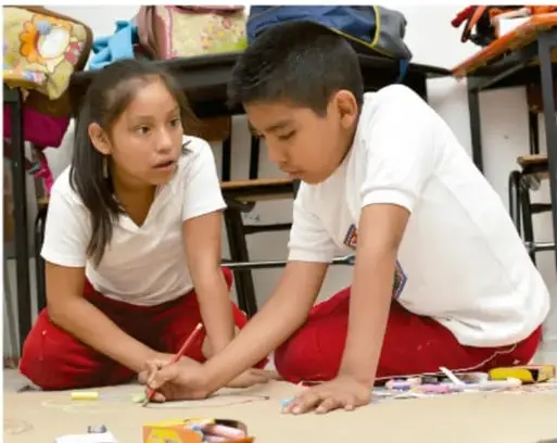 Fotografía de dos estudiantes de primaria (niña y niño) trabajando en el suelo con materiales de arte, pintando o dibujando sobre un papel grande. Están en un aula con mochilas y sillas al fondo.