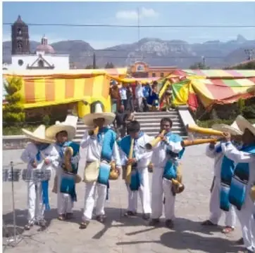 Fotografía de un grupo de danzantes con trajes blancos y azules y sombreros, tocando flautas y tambores frente a una banda de colores en una explanada, con iglesia y montañas al fondo.