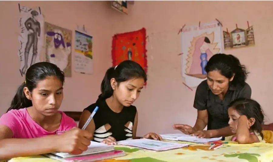 Fotografía de cuatro personas (tres niñas y una adulta) sentadas alrededor de una mesa con manteles de colores, escribiendo en cuadernos; en la pared hay dibujos y murales infantiles colgados con clips. Representa una sesión de trabajo o estudio en comunidad.