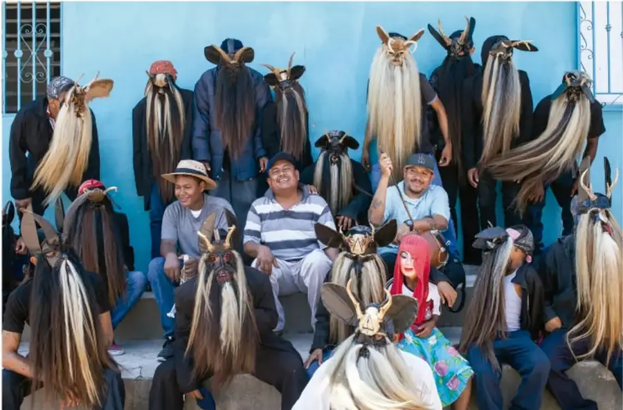 Fotografía de un grupo de personas de una comunidad, varias con máscaras de cabello largo y cuernos (traje tradicional), otras con sombreros, sentadas y de pie frente a un muro azul. Crédito: ©2020 Paola Lagrinos, 1000 DE MÉXICO.