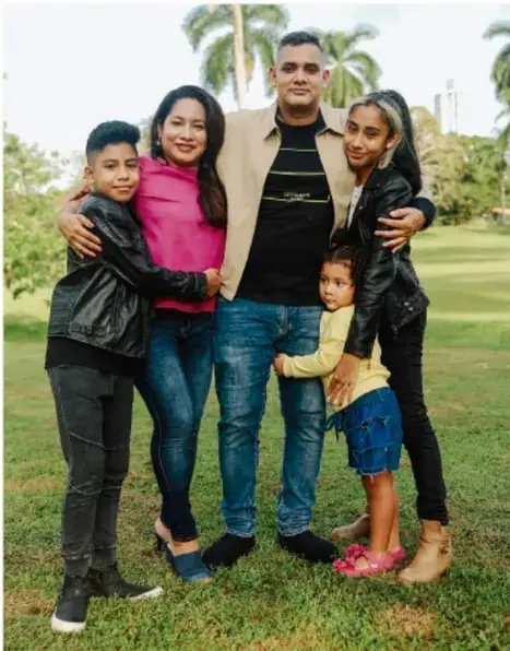 Fotografía de una familia de cinco integrantes (padres, dos adolescentes y una niña pequeña) abrazados en un parque con palmeras al fondo.