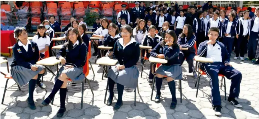 Fotografía de estudiantes de secundaria con uniforme participando en una actividad al aire libre, sentados en pupitres.