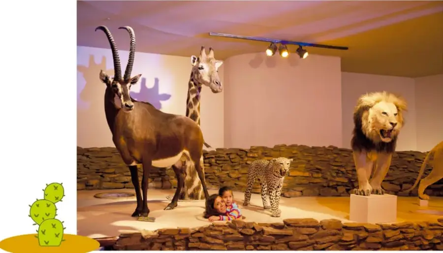 Fotografía de una exhibición museográfica de taxidermia con un antílope de cuernos largos, una jirafa, un leopardo y un león rugiendo sobre un pedestal, junto a dos niñas asomadas detrás de una barrera de piedra.