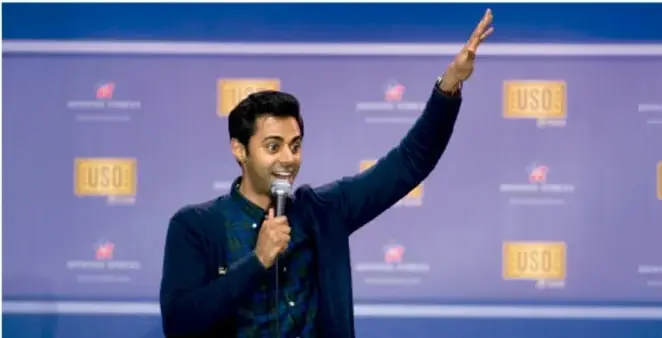 Comediante sobre un escenario con micrófono, brazo en alto, ante un fondo con logotipos (evento de comedia/stand-up).
